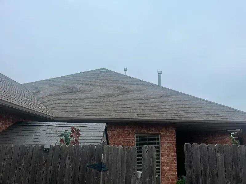 House roof, brick exterior, brown shingles, chimney, wooden fence, overcast sky.