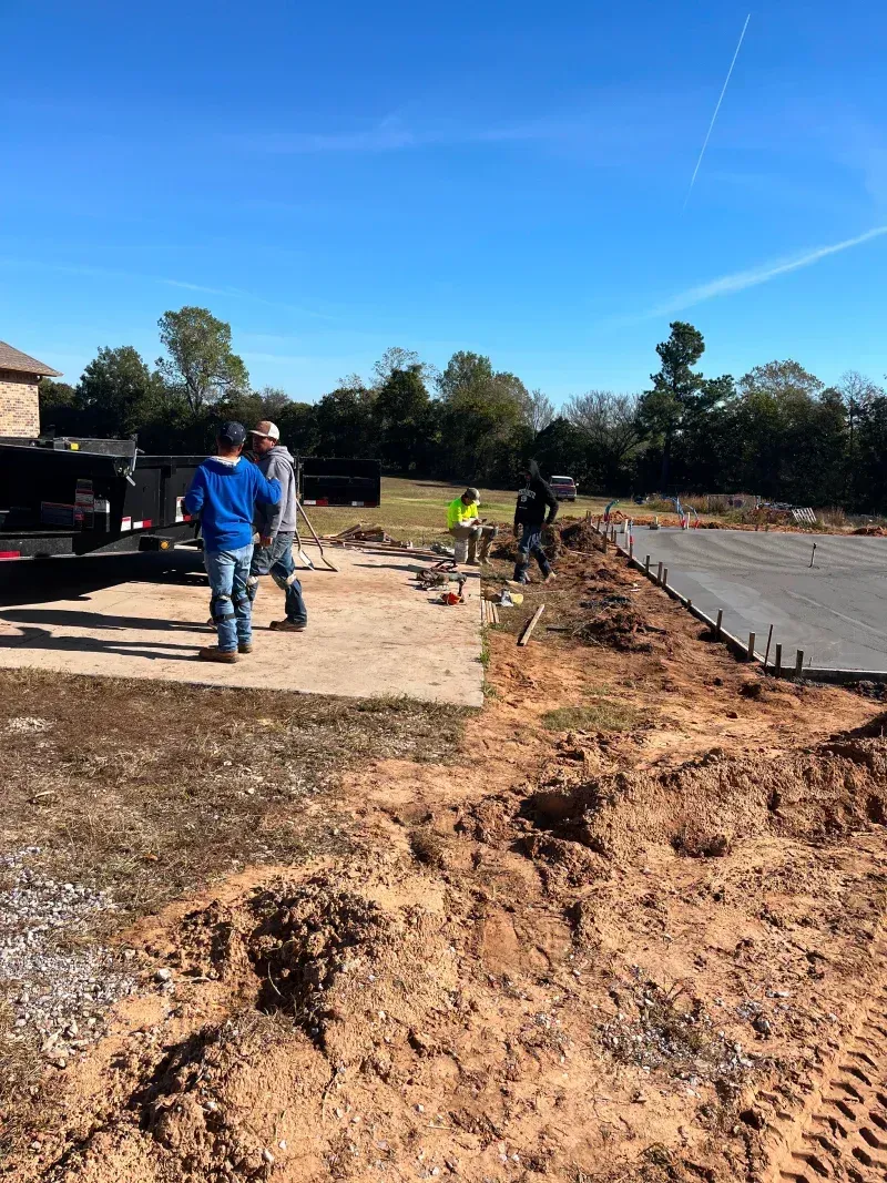 Construction workers on a sunny day building a driveway. Dirt, concrete, and a vehicle are visible.