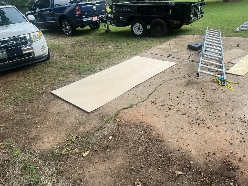 A large sheet of plywood on a driveway. A truck, trailer, and ladder are nearby.