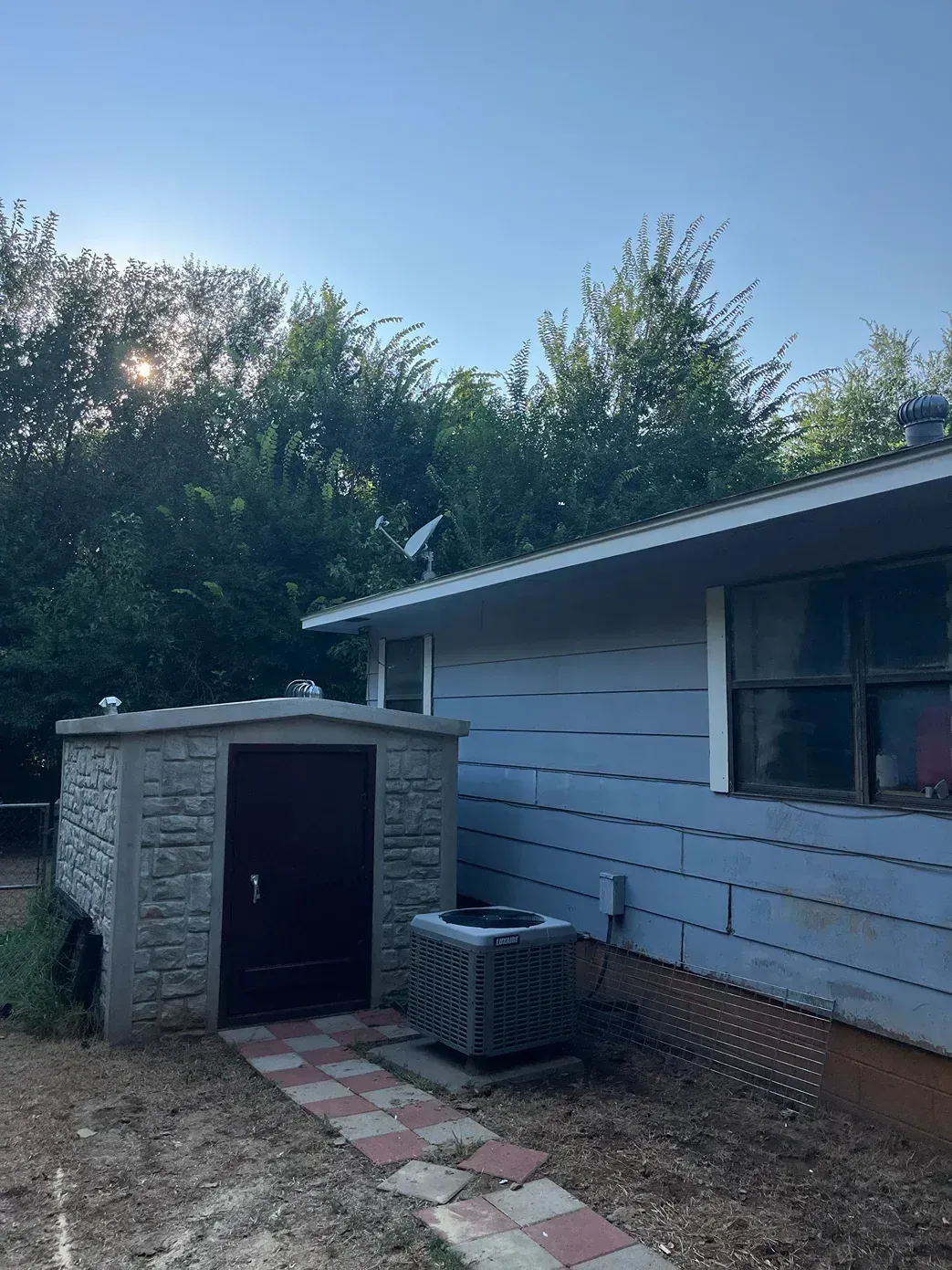 A small stone storage shed next to a light blue house, with an air conditioner unit and a brick pathway.