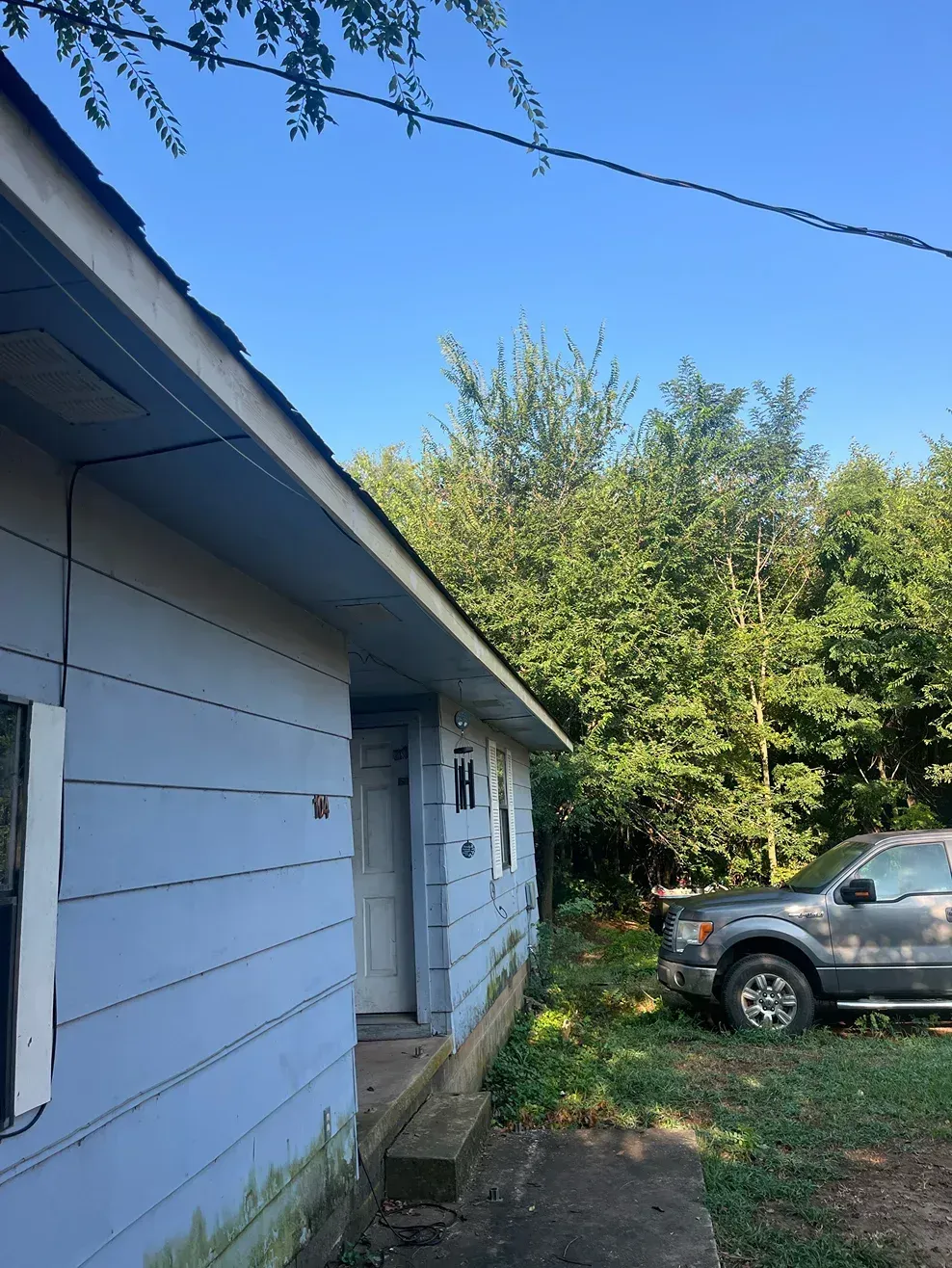 Light blue house with dark roof, overgrown lawn, and parked gray truck.