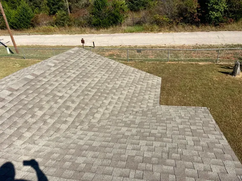 Overhead view of a gray shingle roof with a person taking a photo, surrounded by grass and a road.