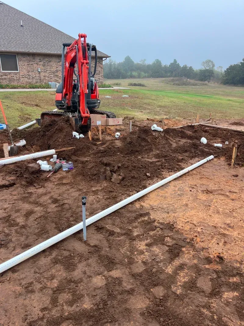 Red excavator on a construction site with exposed pipes and dirt, near a house on a cloudy day.