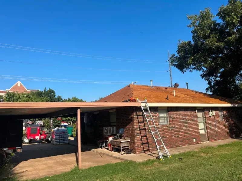 A brick house with a new roof, a carport, and a ladder leaning against it on a sunny day.