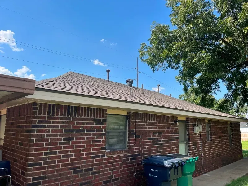 Brick house with brown shingle roof, green trash bins, and a tree under a blue sky.