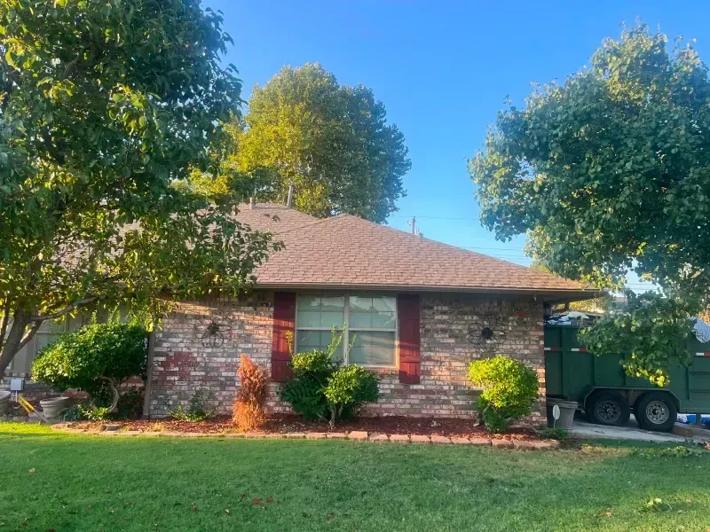 Brick house with brown roof, surrounded by green trees and grass, under a blue sky.