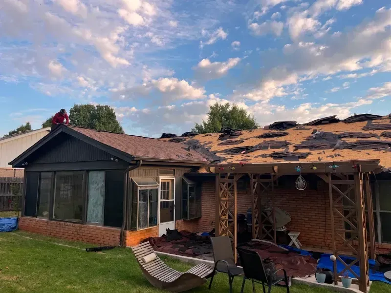 Roof partially torn off a brick house under a cloudy sky. A person works on the roof. Green grass and yard furniture are in view.