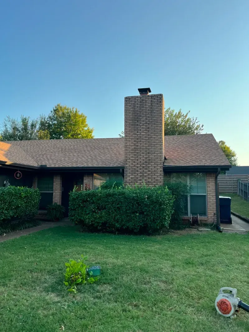House with brown roof, brick chimney, green lawn, bushes, and a leaf blower.