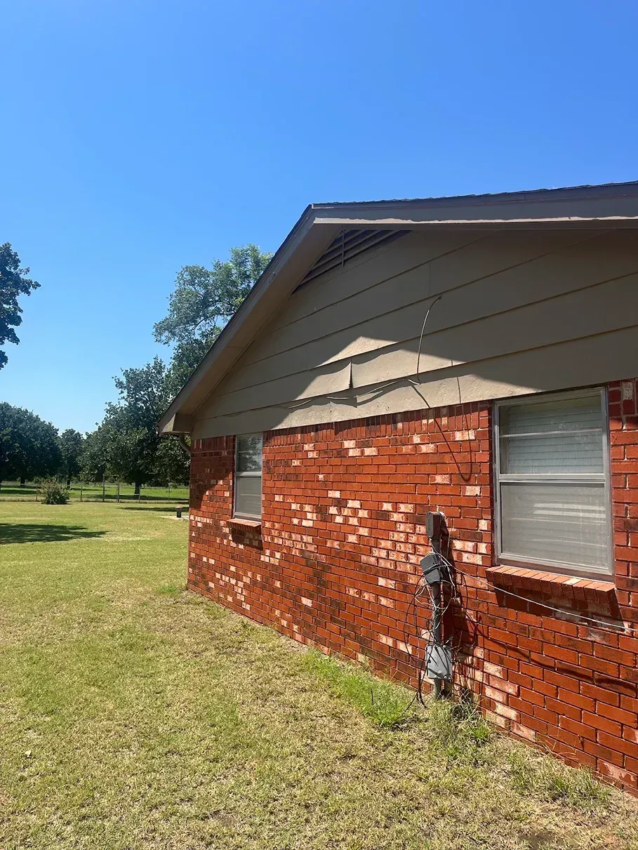Brick house exterior with tan siding and two windows, green lawn, and blue sky.