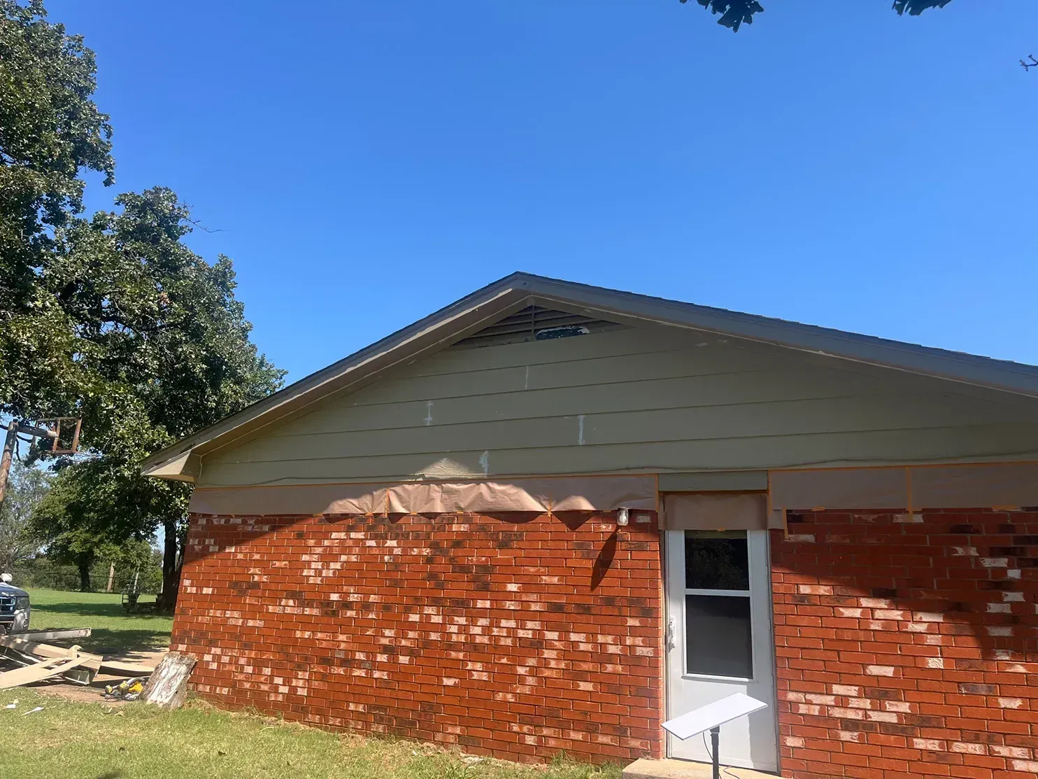 Red brick building with damage under the roof's edge and a white door, blue sky, and a tree in the background.