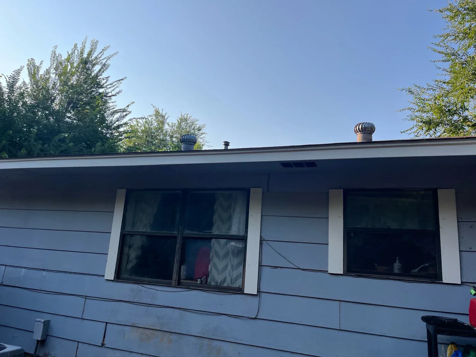 Blue-sided house with two windows and vents on the roof against a blue sky.