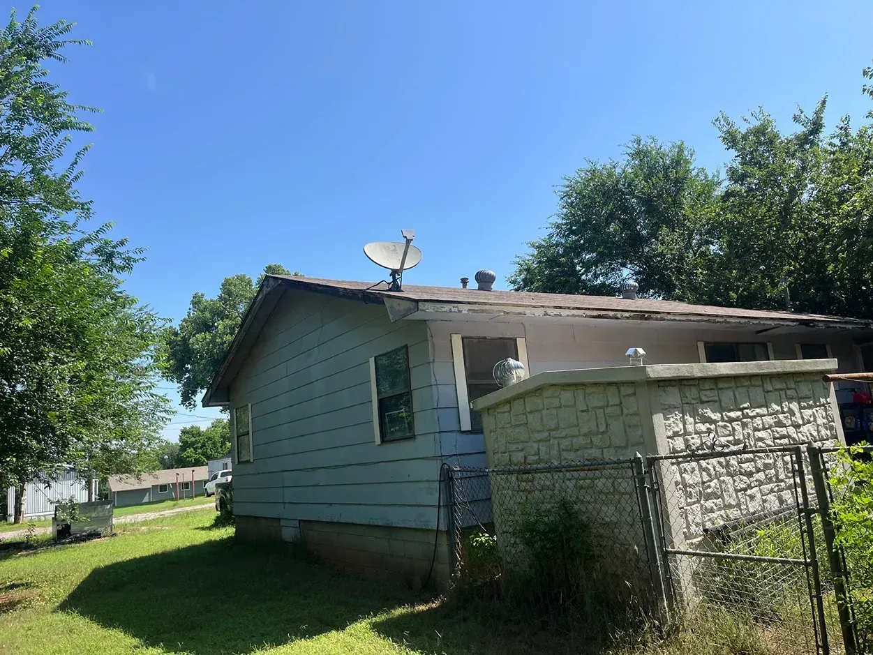 Blue-sided house with damaged roof and satellite dish under a clear, blue sky.