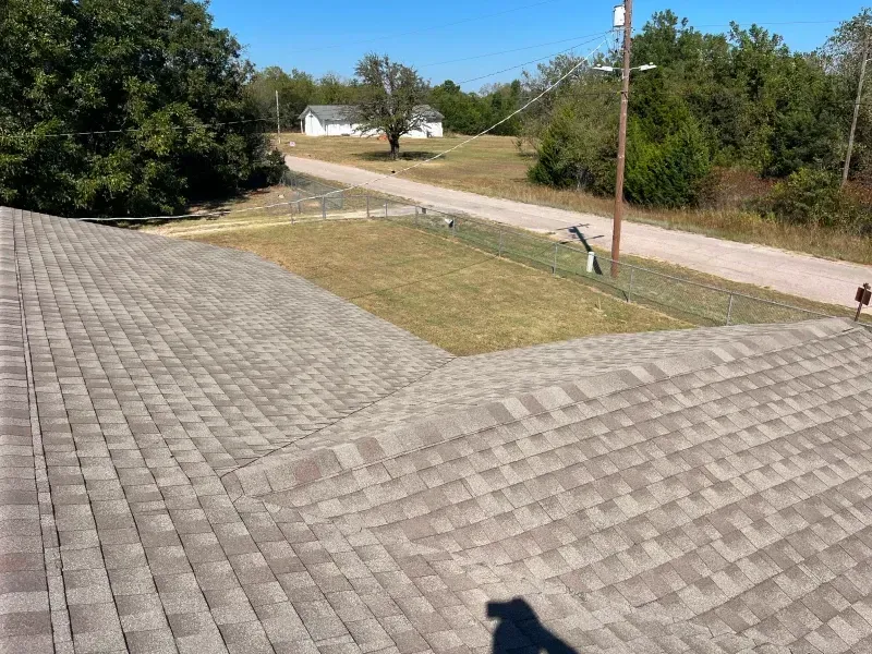 View from a roof, showing asphalt shingles. A road and grassy area lead to distant buildings under a blue sky.