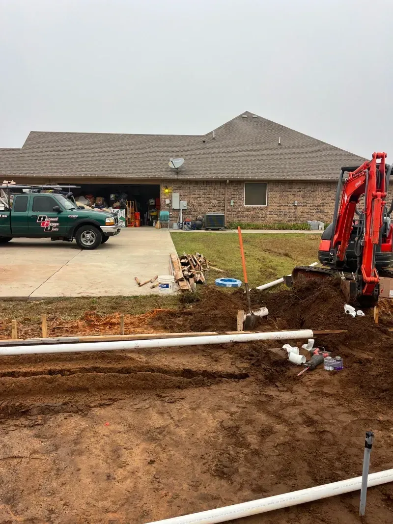 Construction site with a mini-excavator digging near a house; plumbing pipes laid in a trench.