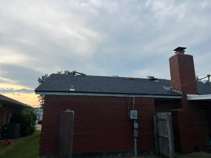 Brick house with a newly shingled roof, a chimney, and tools visible on the roof against a cloudy sky.