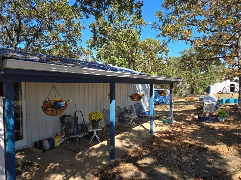 Small white building with a covered patio, hanging baskets, and chairs. Blue accents and a yard with trees.