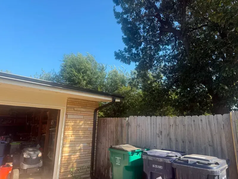 Garage with dark gutters and a wooden fence; trash cans in the yard, tree in the background under a blue sky.