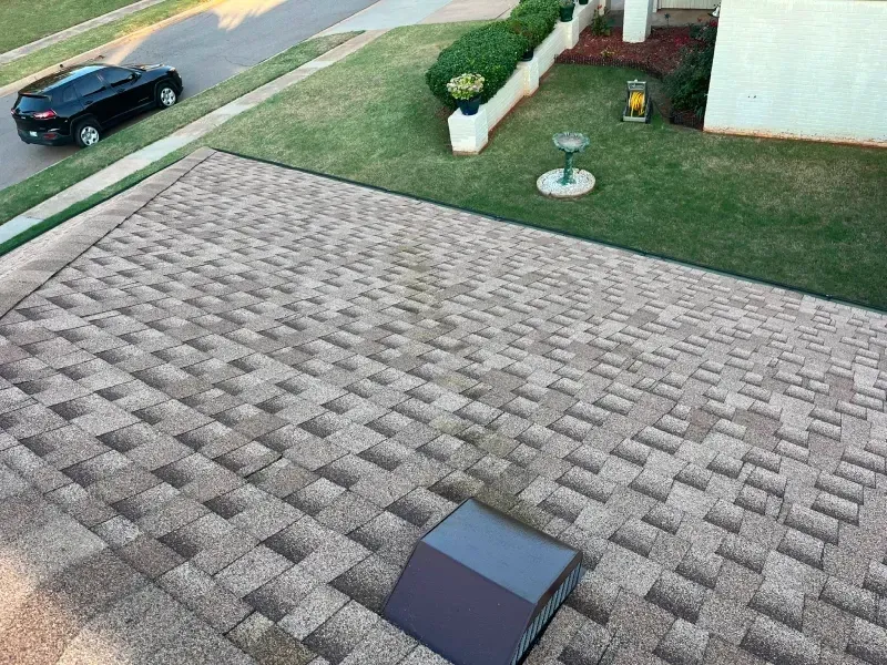 View from a roof, showing asphalt shingles in shades of brown and a small vent. A black car sits on the street below.