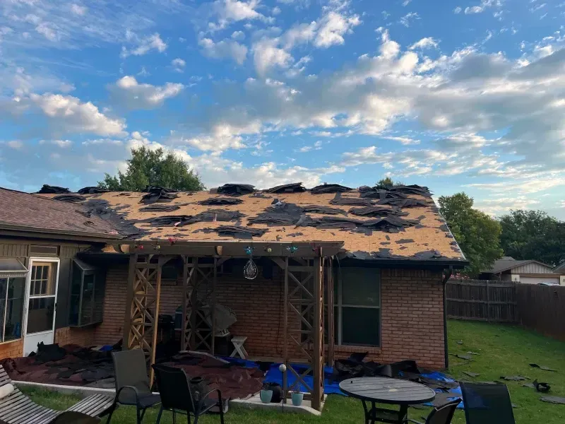Roof with missing shingles, debris on the ground, brick house, cloudy sky, backyard setting.