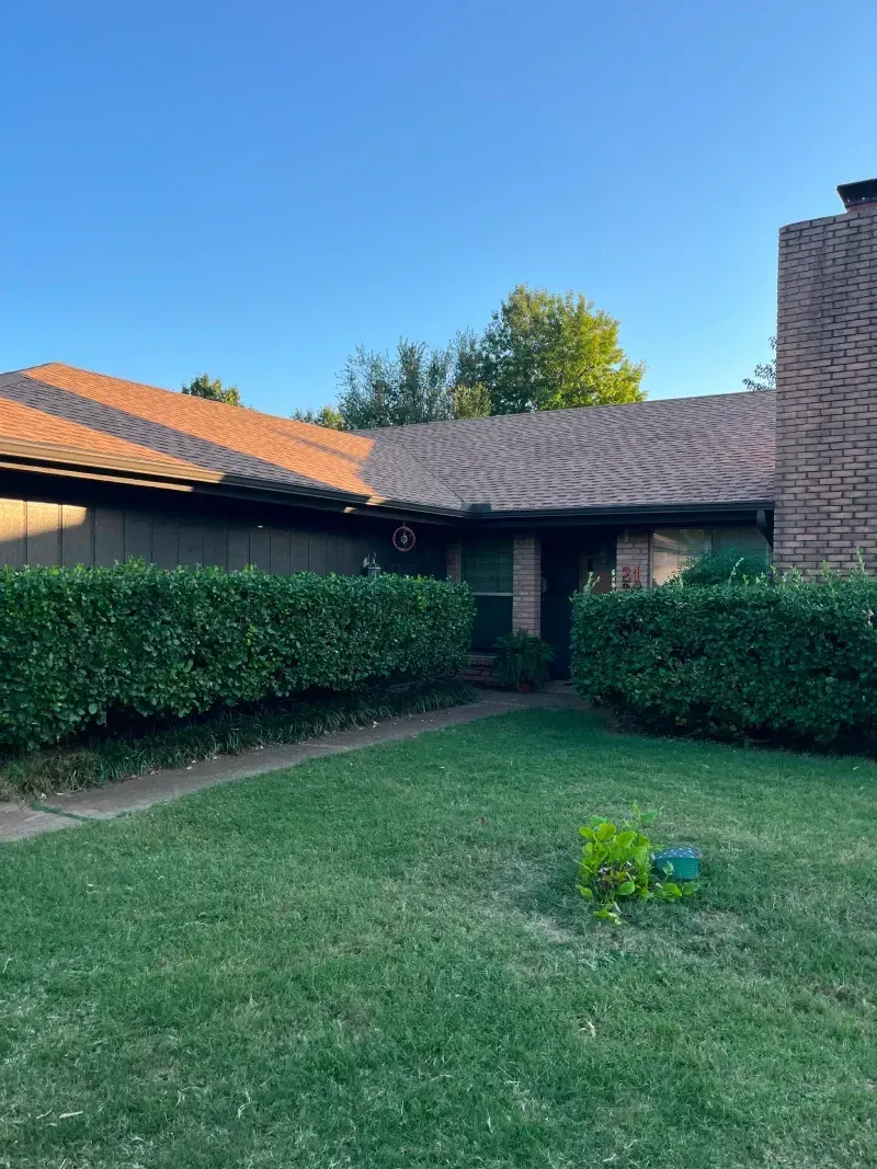 Brown-roofed house with brick chimney, dark siding, and trimmed green hedges in front of a green lawn under a blue sky.