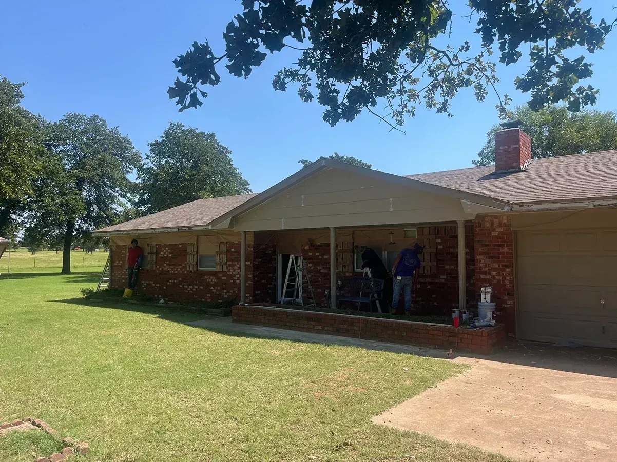 Brick house with workers on porch; green lawn, blue sky, trees.
