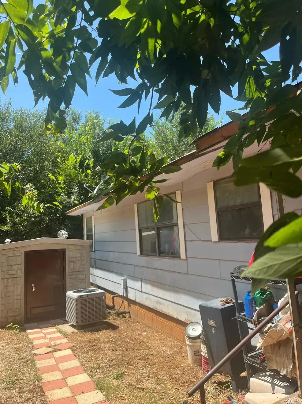 Side of a light blue house with two windows and a small shed in a yard, under tree branches.
