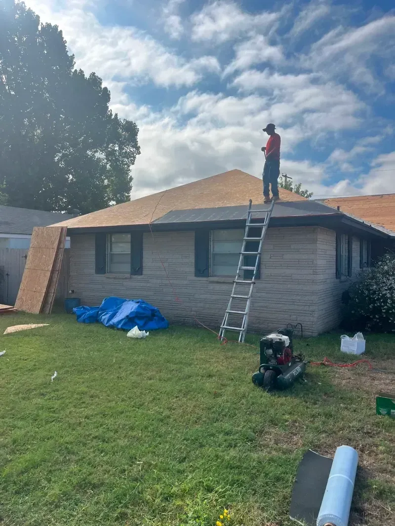 Person on a ladder repairing a brown shingle roof on a brick house; green grass, blue sky.
