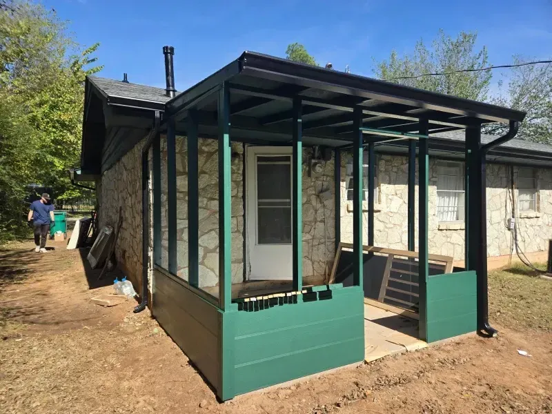 Green screened-in porch addition attached to a stone-walled house, under a clear blue sky.
