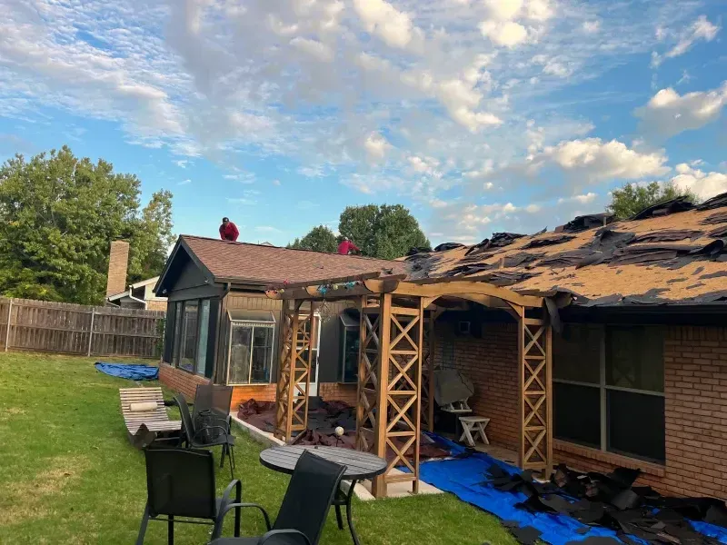 Roof damage on a house with workers. Backyard scene with torn shingles, wooden pergola, and blue tarp.