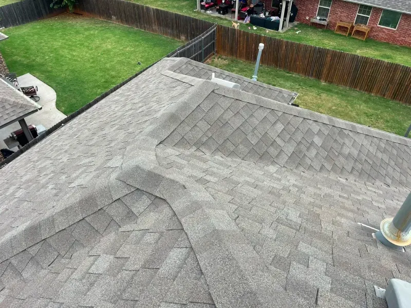 Gray shingle roof, angled view. Chimney, vents, and edges visible. Green lawn and fences in background.