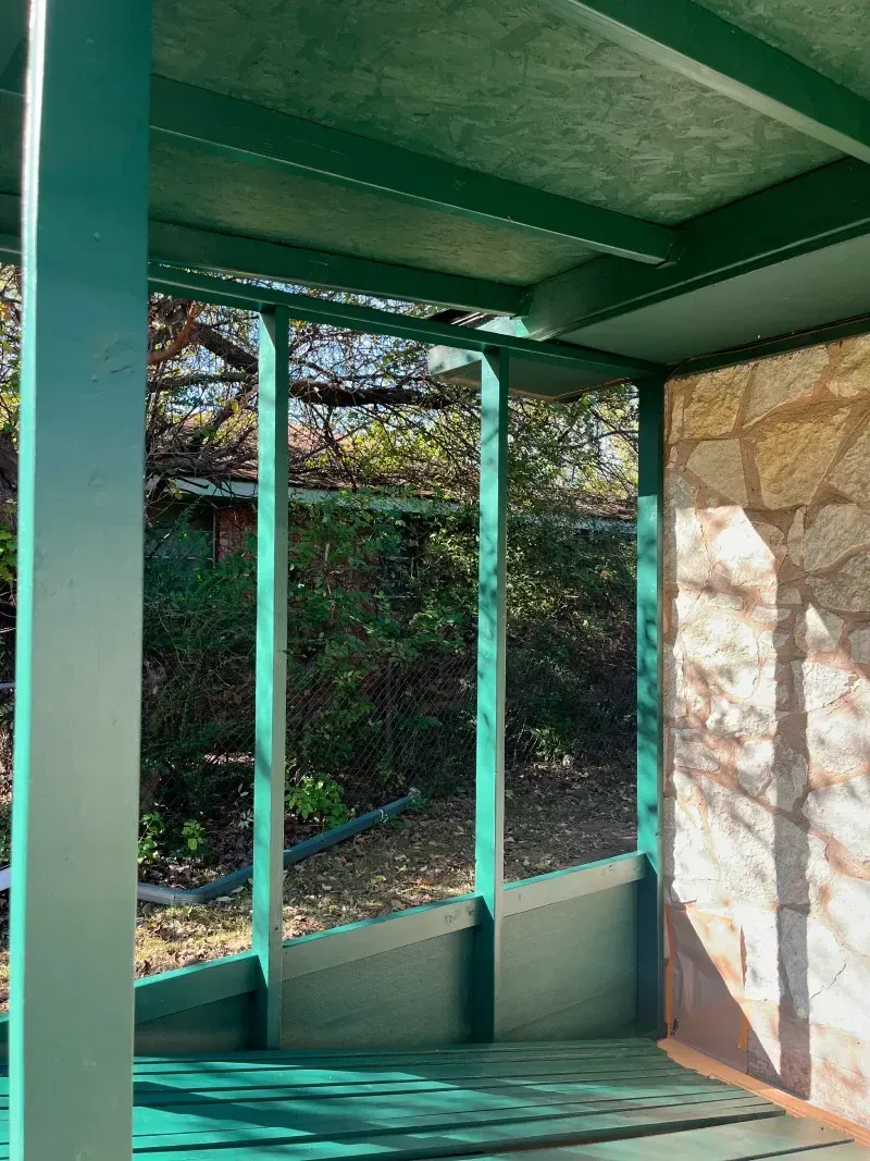 Green screened-in porch with view of trees and stone wall.