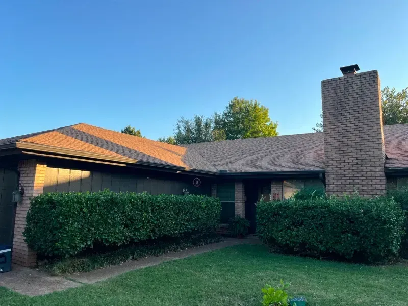 A one-story house with a brown roof and a brick chimney, green bushes in front.