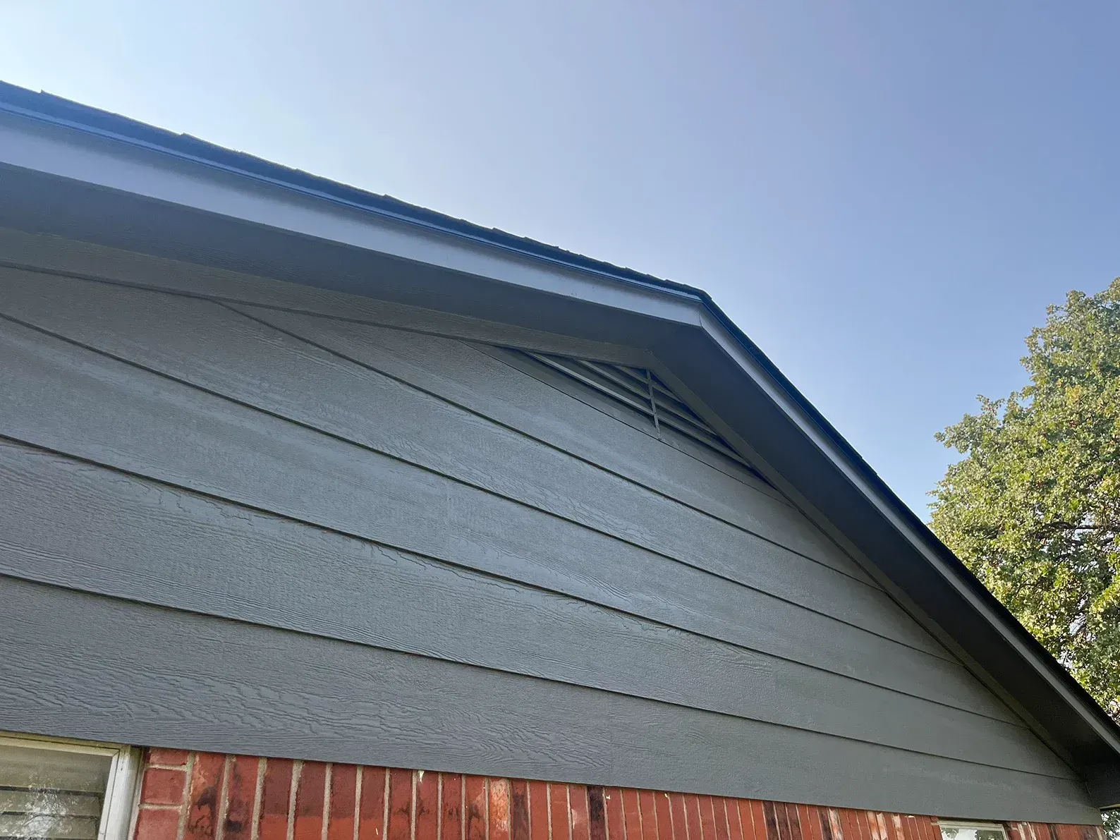 Gray siding and roof of a house against a blue sky, with a brick base visible.