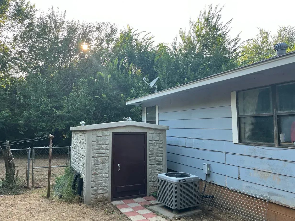 Gray shed with a dark door next to a pale blue house with trees in the background, a satellite dish, and air conditioner.