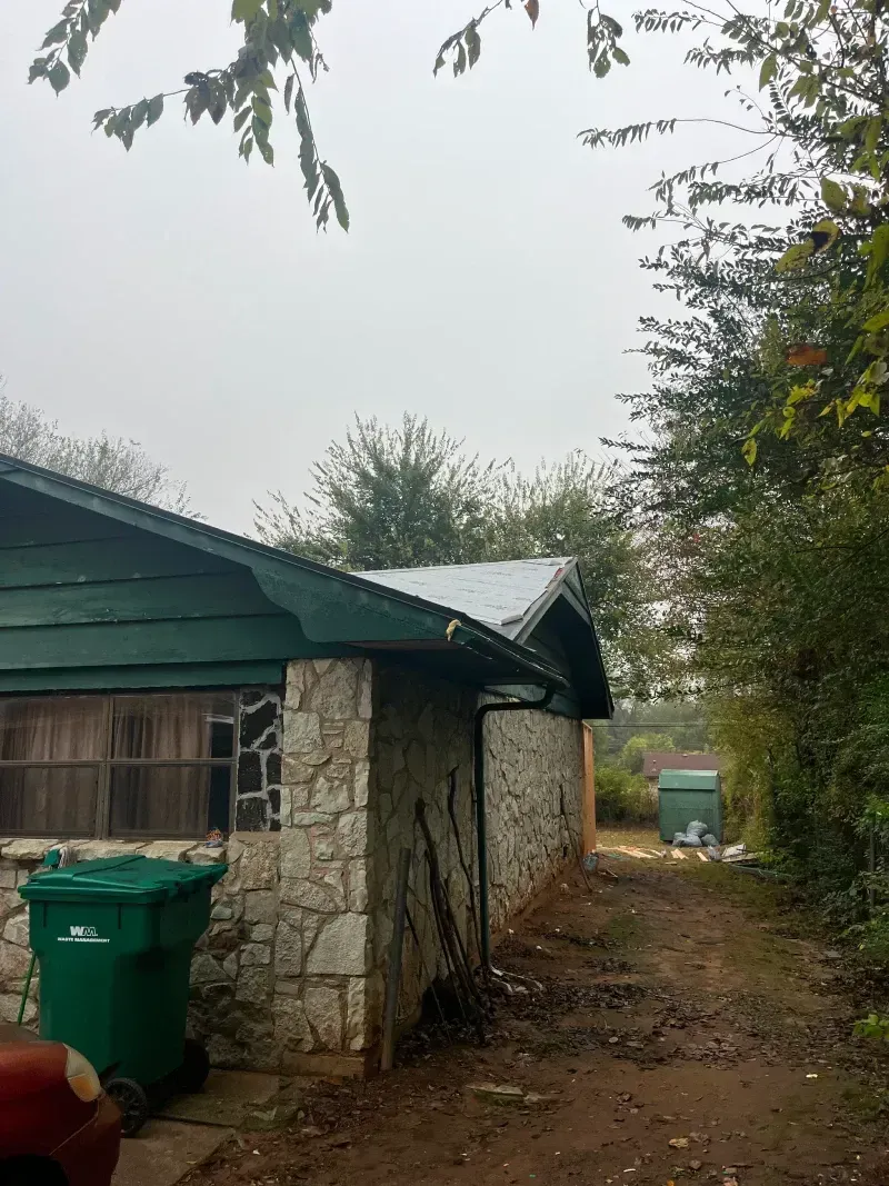 Stone and wood building with green trim, green trash can, muddy path, overcast sky.