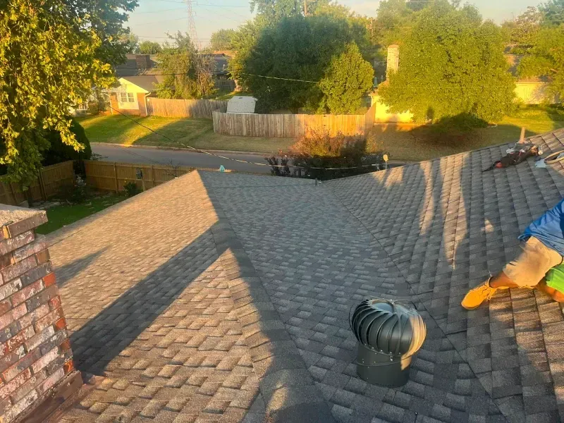 Rooftop view of a home with new shingles and a spinning vent. Houses and trees in the background under a sunny sky.