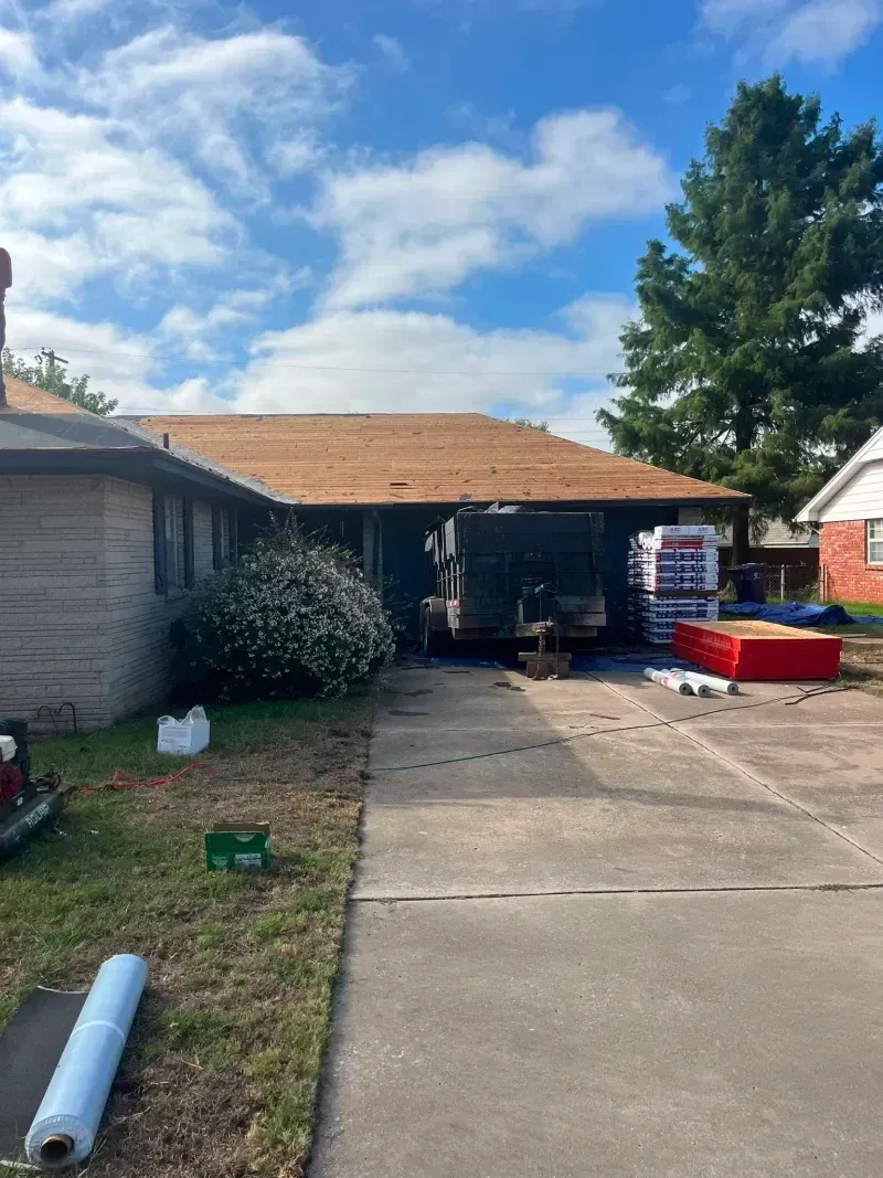 House with brown roof under construction, trailer parked in driveway with roofing supplies.