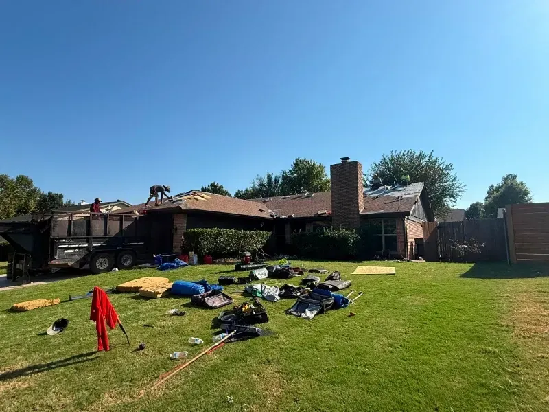 House roof replacement in progress; workers on roof, debris on lawn, blue sky.