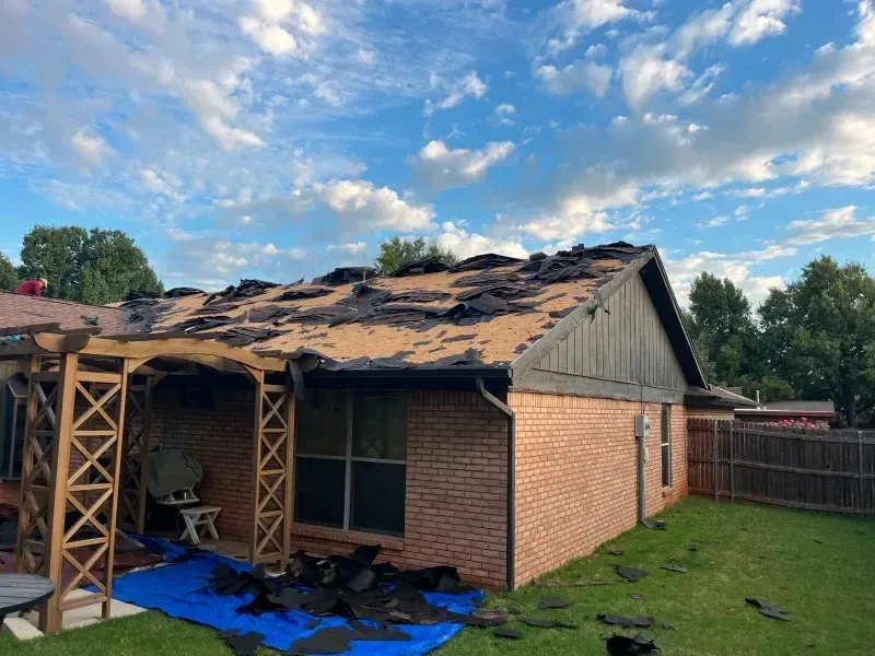 House with torn-off roof shingles, debris on ground, blue tarp, brick exterior, and pergola; cloudy sky.