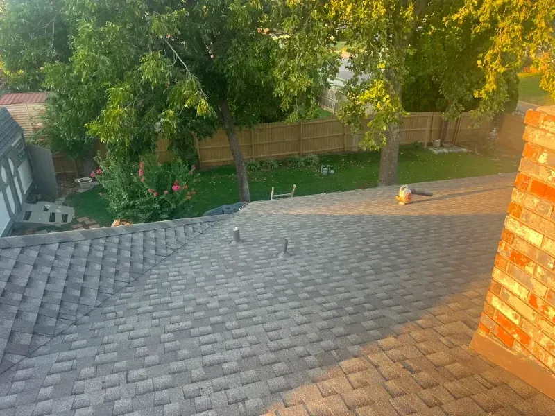 Overhead view of a house roof with a brick chimney, overlooking a green yard and trees.