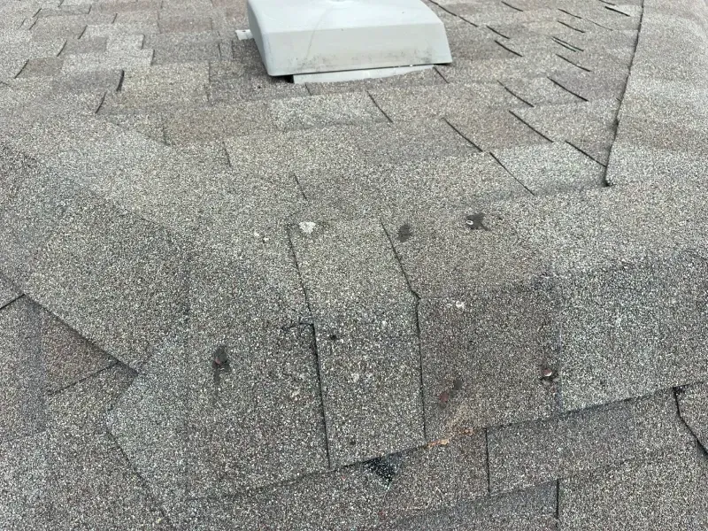 Close-up of a weathered shingle roof with a vent. The shingles are gray with granular texture and some missing nails.