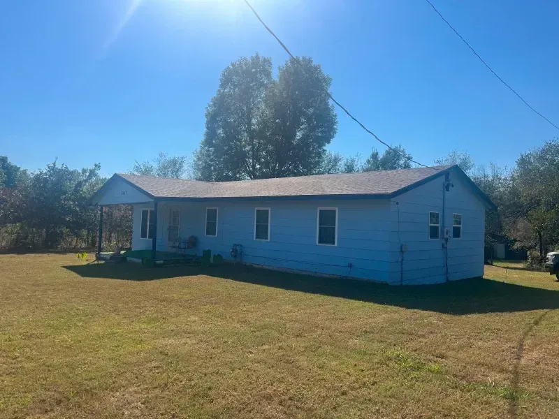 Blue house with a porch and brown roof, set in a grassy yard under a blue sky.