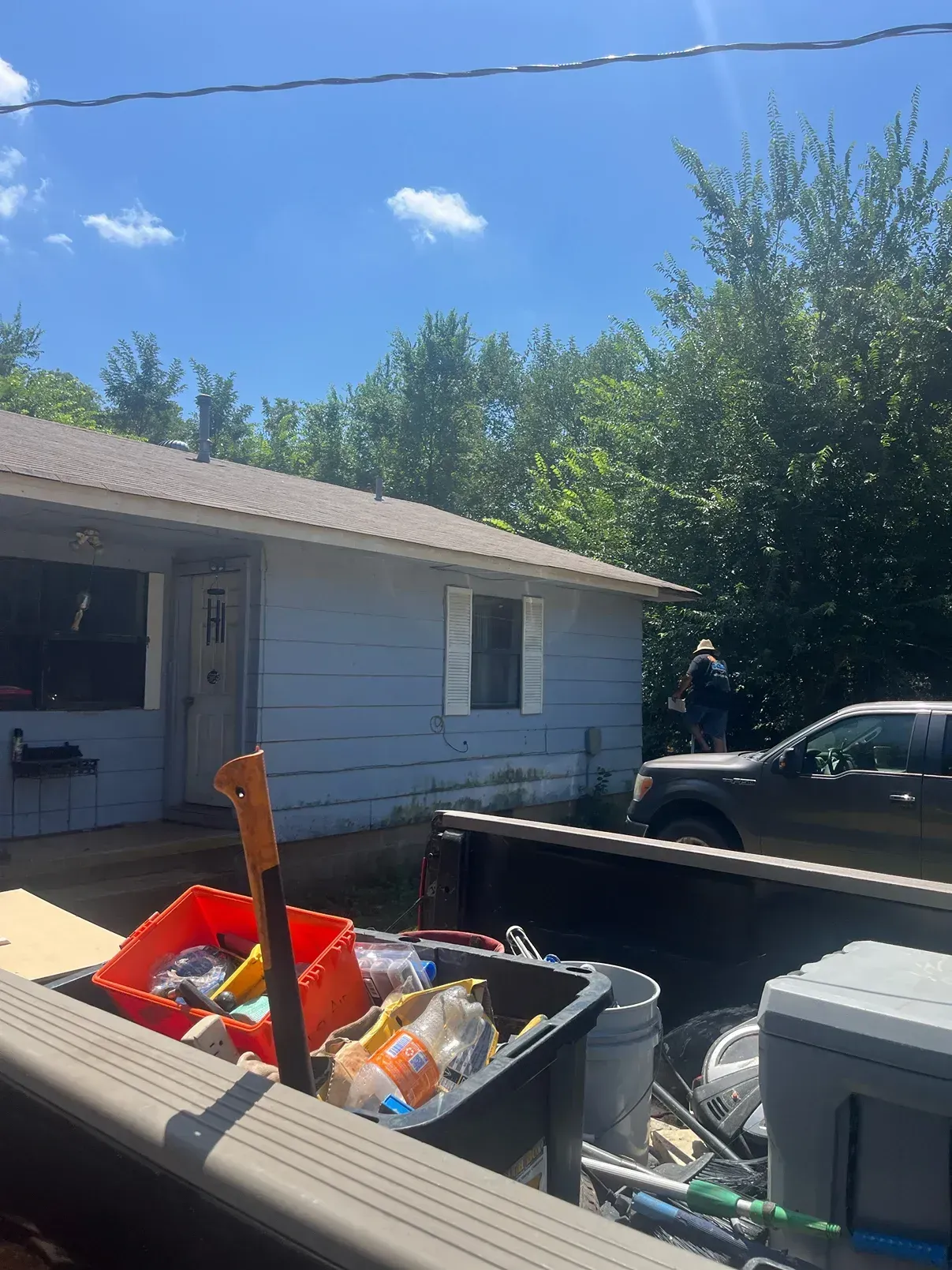 Blue house exterior, truck bed filled with items. Person standing by car, trees in background, sunny day.
