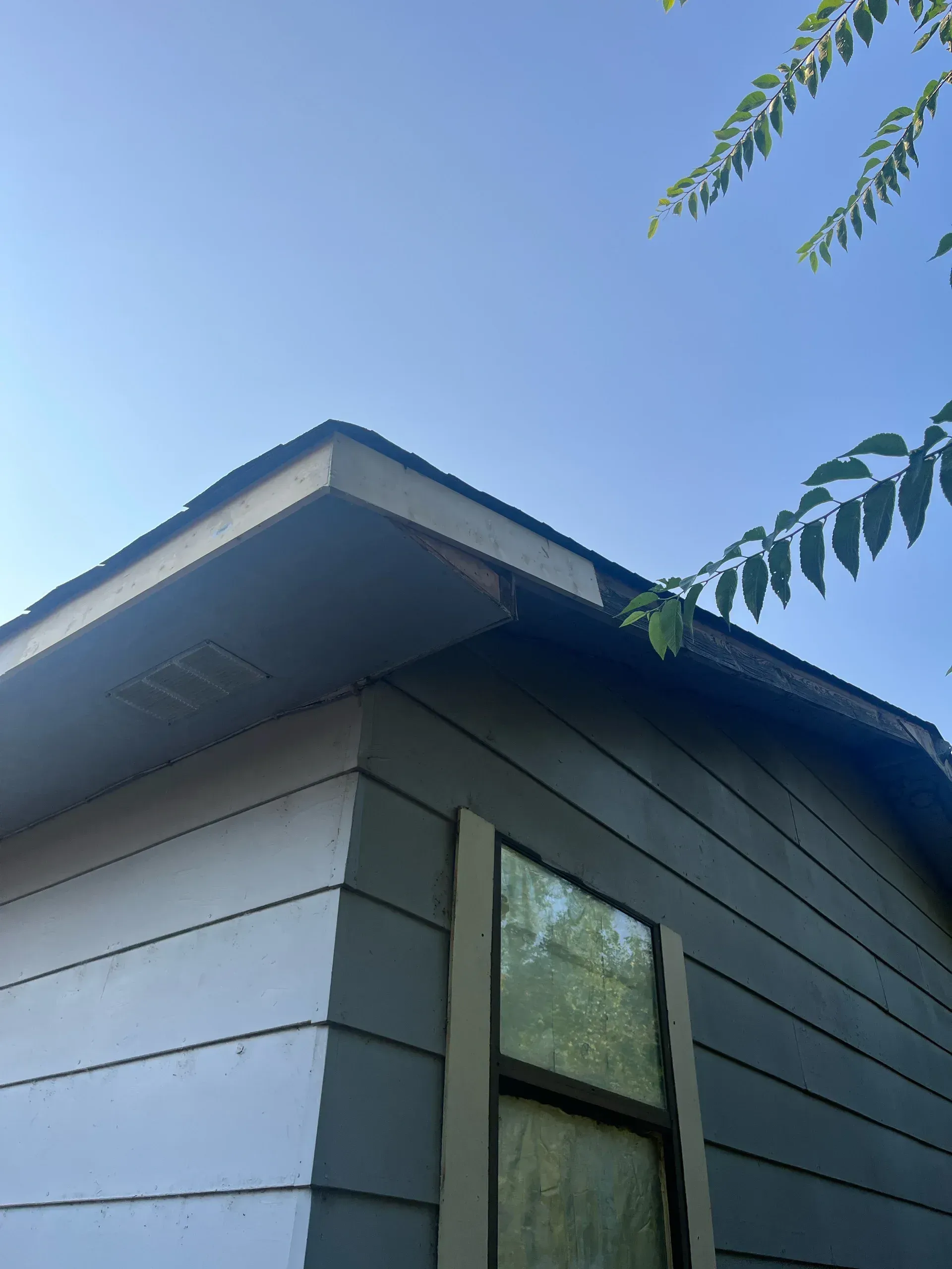 Corner of a house with gray siding, a window, and a damaged roofline against a blue sky.