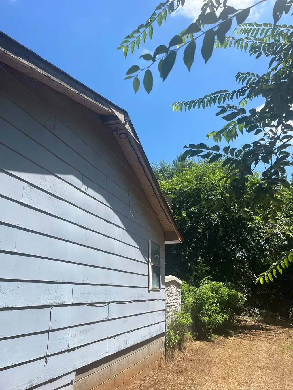 Side view of a blue-sided house with a tan roof against a blue sky with trees.