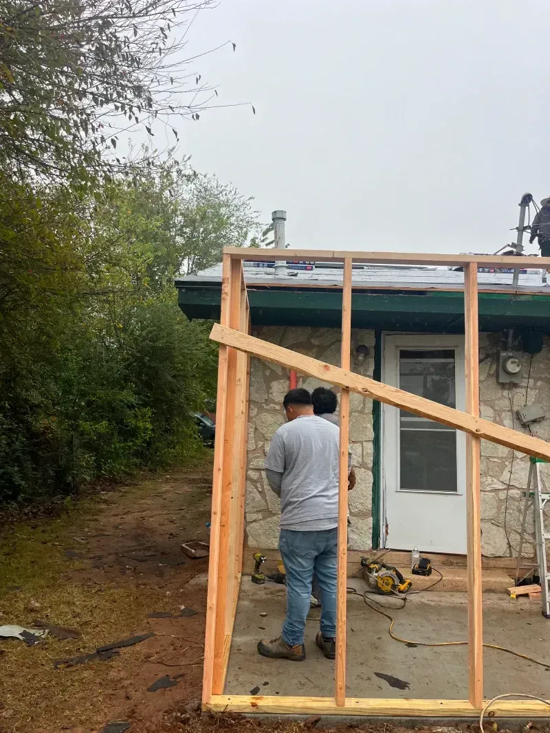Two people framing a wooden structure against a stone building with a doorway, overcast day.