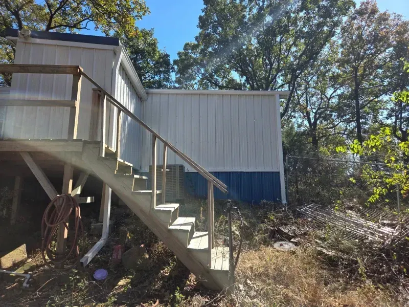 Exterior of a building with blue and white siding. Wooden stairs lead up to a deck. Trees surround.