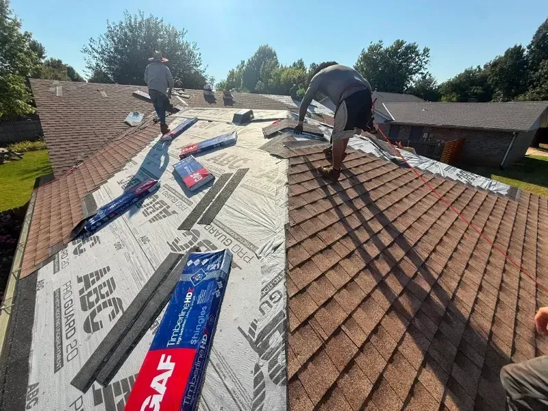 Roofers working on a brown shingled roof, some shingles removed, materials and tools present.