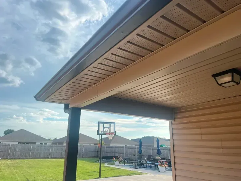 View of a brown-sided house with a covered patio, basketball hoop, and lawn under a cloudy sky.