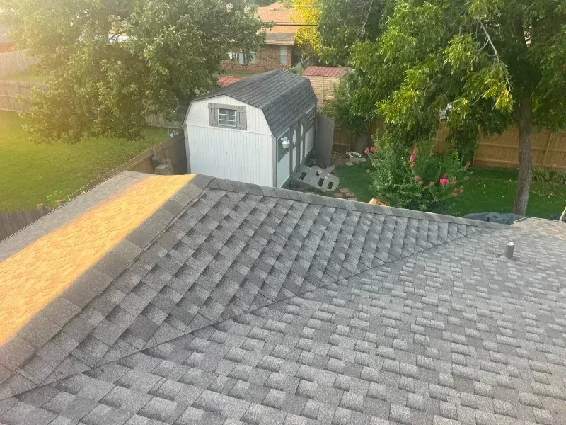 View from a roof of a suburban house. Gray shingles, shed in the yard, trees and green grass.